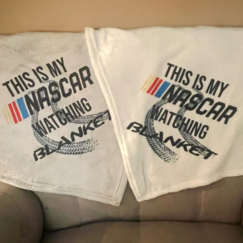 May include: Two white fleece blankets with the text "This is my NASCAR watching blanket" printed on them. The blankets feature a tyre tread design and a red, white and blue chequered flag.