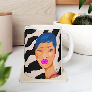 May include: A white ceramic mug featuring a colourful illustration of a person with short blue hair and bright pink lips. The background of the design consists of a black and beige zebra stripe pattern. The artist's signature is visible on the lower right.