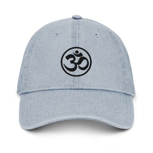 Könnte beinhalten: Hellblaue Jeans-Baseballkappe mit einem schwarzen Om-Symbol in einem Kreis auf der Vorderseite. Die Kappe hat einen gebogenen Schirm und eine strukturierte Krone. Ein lässiges Accessoire.