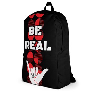 May include: Black backpack with the words "BE REAL" in white block letters. The backpack features a red and white graphic design and a white hand making a shaka sign.