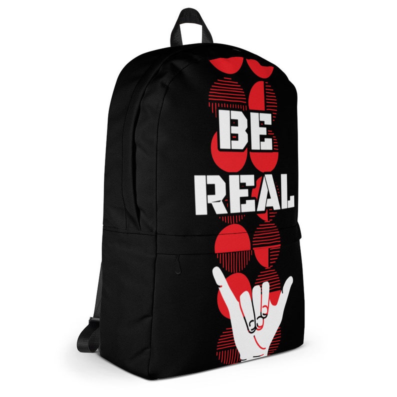 May include: Black backpack featuring the text "BE REAL" in white, red circular designs, and a white hand gesture. The bag has a front pocket, a top handle, and adjustable shoulder straps. Ideal for carrying books and essentials.