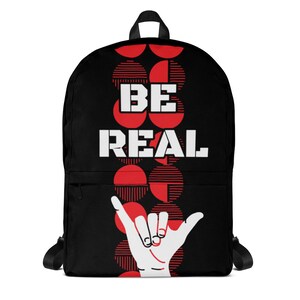 May include: Black backpack featuring the words "BE REAL" in white and a white hand gesture. The background has a pattern of red circles. The backpack has black straps and a front pocket.