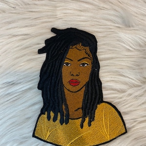 May include: Embroidered patch featuring a woman with long black dreadlocks, wearing a yellow shirt. Her eyes are brown, and she has red lips.