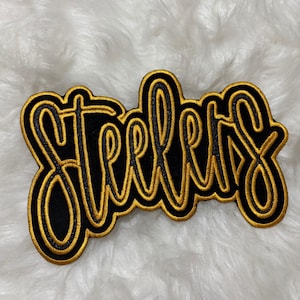 May include: Black and gold embroidered patch with the word "Steelers" in a cursive font. The lettering is outlined in gold thread, with a black interior. The patch is likely for a jacket, bag, or other apparel.