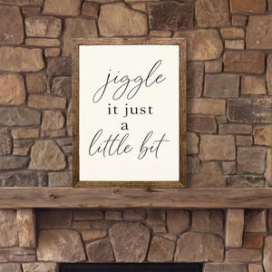 May include: A rectangular wall sign with a dark wood frame and a white background featuring the black text "jiggle it just a little bit" in a combination of cursive and serif fonts, mounted on a stone fireplace above a hearth.