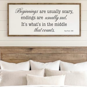 May include: A bedroom scene with a wooden headboard, two windows, and a bed with white bedding. A framed sign above the bed reads "Beginnings are usually scary, endings are usually sad. It's what's in the middle that counts." Two lamps and nightstands flank the bed.