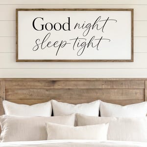 May include: A rectangular wall sign with a dark wood frame and a white background featuring the black cursive text Good night sleep tight. The sign is centered above a wooden headboard in a bedroom setting with two windows and bedside lamps.