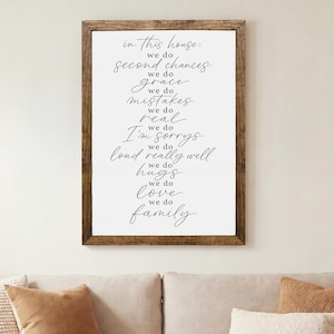 May include: A rectangular wall sign with a dark wood frame featuring black cursive text on a white background that reads: in this house we do second chances we do grace we do mistakes we do real we do I'm sorrys we do loud really well we do hugs we do love we do family.