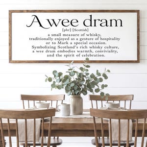 May include: A rectangular wall sign with a white background and a dark wood frame, featuring black printed text that defines the phrase A wee dram as a small measure of whisky, symbolizing Scotland's rich culture, warmth, and celebration.