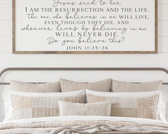 May include: A bedroom scene featuring a bed with a neutral-toned quilt and pillows. Above the bed is a framed sign with a religious quote. The room has a farmhouse aesthetic with white walls, bedside tables, and lamps.
