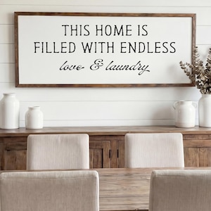 May include: A dining room scene featuring a wooden table and chairs. A framed sign on the wall reads "THIS HOME IS FILLED WITH ENDLESS love & laundry." Decorative vases and dried flowers add to the room's aesthetic.