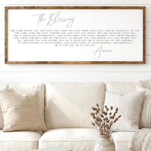May include: A framed wooden sign with the text "The Blessing" in elegant script. The sign features a prayer written in a classic font. The sign is displayed above a white sofa with neutral-colored pillows and a wooden coffee table.
