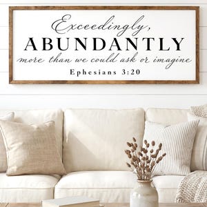May include: A framed wooden sign with the text "Exceedingly, Abundantly more than we could ask or imagine. Ephesians 3:20" in black script. The sign hangs above a white sofa with beige pillows. The words "FULLY CUSTOMIZABLE - SIZES, COLORS" are at the bottom.