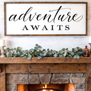 May include: A rectangular wall sign with a dark wood frame and a white background featuring the black text adventure AWAITS in a combination of cursive and block lettering. The sign is mounted horizontally above a wooden fireplace mantel.