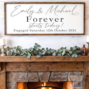 May include: A rectangular white wooden sign with a dark brown frame featuring black text that reads Emily & Michael, Forever starts today!, and Engaged Saturday 12th October 2024. The sign is displayed horizontally above a rustic wooden fireplace mantel.