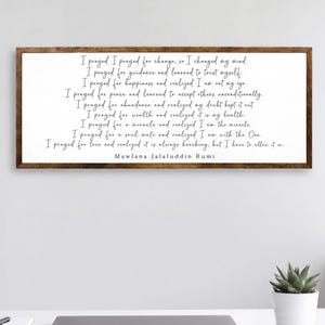 May include: A framed wall art piece with a white background and dark brown frame. The artwork features a quote by Mawlana Jalaluddin Rumi in black script. The quote is a series of "I prayed" statements, reflecting on personal growth and realization.