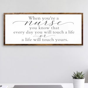May include: A rectangular wall sign with a dark wood frame and a white background featuring black text that reads: When you're a nurse you know that every day you will touch a life or a life will touch yours. The sign has a clean, simple design.