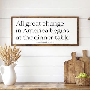 May include: A framed sign with the quote "All great change in America begins at the dinner table" by Ronald Reagan. The sign is above a wooden table with a white pitcher of wheat, cutting boards, and a basket of green apples.