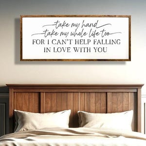 May include: A wooden framed sign with the text "take my hand take my whole life too FOR I CAN'T HELP FALLING IN LOVE WITH YOU" hangs above a wooden bed. The sign is white with black lettering. The bed is brown with white bedding.