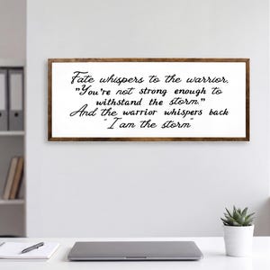 May include: A rectangular wall sign with a dark wood frame and a white background featuring black cursive text that reads: Fate whispers to the warrior, You're not strong enough to withstand the storm. And the warrior whispers back I am the storm.