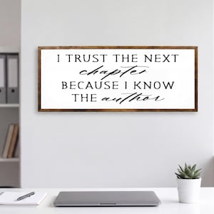 May include: A rectangular wooden sign with the text "I TRUST THE NEXT chapter BECAUSE I KNOW THE author" in black script. The sign is mounted on a white wall above a desk with a laptop, notepad, and pen.