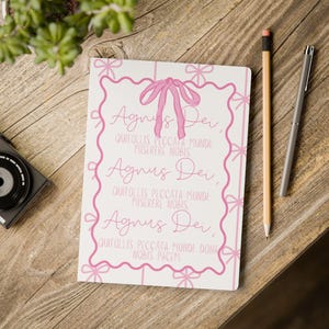 Könnte beinhalten: Ein weißes Notizbuch mit rosa Text und einem rosa Schleifendesign. Der Text lautet "Agnus Dei" und andere lateinische Sätze. Ein Bleistift und ein Stift liegen neben dem Notizbuch auf einer Holzoberfläche. Eine Kamera und eine Pflanze sind ebenfalls sichtbar.