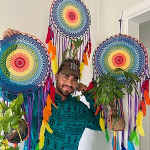 May include: Three vibrant dream catchers with rainbow-hued centres and colourful feather embellishments. Each dream catcher features hanging ribbons and a small plant. The dream catchers are suspended from the ceiling.