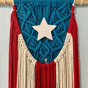 May include: A macrame wall hanging featuring the flag of Puerto Rico. The design includes a blue field with a white star, red stripes, and white stripes. The piece is suspended from a wooden dowel with a knotted cord.