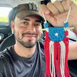 Puede incluir: Un tapiz de macramé con la bandera de Puerto Rico. El diseño incluye una sección superior azul con una estrella blanca, rayas rojas y blancas y una clavija de madera. La pieza se cuelga con un cordón blanco.