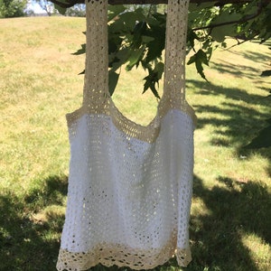 May include: A white crochet tank top with a cream colored crocheted trim. The tank top is hanging from a tree branch.