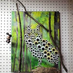 May include: A green and yellow owl painting made with googly eyes. The owl is perched on a brown branch with a green and brown forest background.