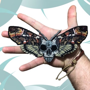 Könnte beinhalten: Ein gestickter Aufnäher eines Totenkopfschwärmers mit braunen, orangefarbenen und weißen Flügeln.