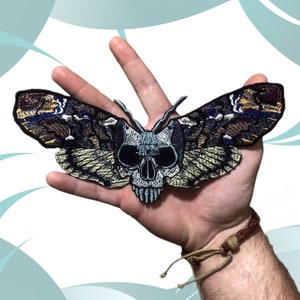 Könnte beinhalten: Ein gestickter Patch in Schwarzweiß, der eine Motte mit einem Schädel als Kopf zeigt. Die Motte hat braune, blaue und gelbe Flügel.