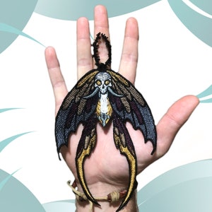 Puede incluir: Un parche bordado de color negro y dorado de una polilla con una calavera y colmillos. La polilla tiene alas grandes con detalles intrincados.