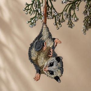 May include: Embroidered opossum ornament hanging from a brown stem with green and silver foliage. The opossum has gray, tan, and black details, with pink paws and a detailed stitched design. A unique holiday decoration.