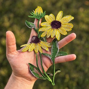 May include: Embroidered floral patches featuring yellow sunflowers with dark purple centers and green leaves. The patches are detailed with intricate stitching and are displayed against a blurred green background.