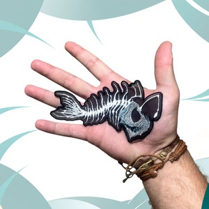 Puede incluir: Un parche bordado en blanco y negro que representa un esqueleto de pez. El parche está en la mano de una persona.