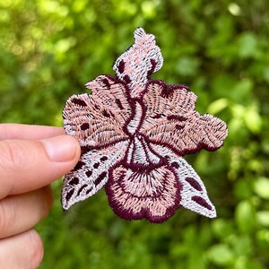 May include: Embroidered floral patch in shades of pink, white, and burgundy. The patch features a detailed orchid design with intricate stitching. The patch is held in a hand against a blurred green background.