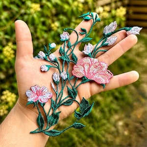 Puede incluir: Aplicación floral bordada con flores rosas y blancas, tallos y hojas verdes. La aplicación tiene un diseño detallado y texturizado, ideal para embellecer ropa o manualidades. Las flores están en plena floración.