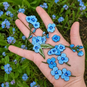 May include: Embroidered blue forget-me-not flower patches with green leaves and stems. The patches are in various sizes and shapes, perfect for adding to clothing or accessories. The patches are displayed on a hand with a background of forget-me-nots.