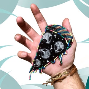 Puede incluir: Un parche bordado en blanco y negro de una rebanada de pizza con glaseado de arcoíris que gotea. La pizza está cubierta con tres caras de extraterrestres y estrellas.