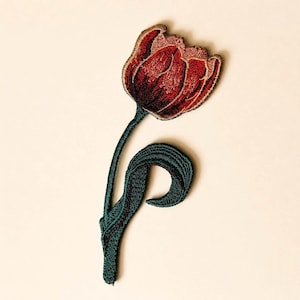 Op de afbeelding: Geborduurde tulp patch. De bloem is dieprood met een gouden omtrek, de stengel en het blad zijn groen. Deze patch is waarschijnlijk voor kleding of accessoires. Het ontwerp is gedetailleerd en gestructureerd, wat een bloemig accent toevoegt.