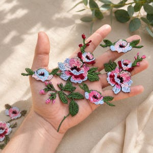 Puede incluir: Apliques florales bordados en rosa, rojo y azul con hojas verdes. Los apliques presentan costuras detalladas y están dispuestos sobre un fondo neutro. Estos parches decorativos son ideales para añadir un toque floral a la ropa o accesorios.