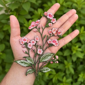 Könnte beinhalten: Gestickter Blumenaufnäher mit rosa Blüten mit gelben Zentren und grünen Blättern. Der Aufnäher hat ein detailliertes Design mit schwarzer Umrandung. Der Aufnäher wird in einer Hand vor einem grünen, belaubten Hintergrund gehalten.