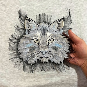 May include: A close-up of a black and white embroidered lynx with a fluffy, fringed border. The lynx has blue eyes and a light gray muzzle.