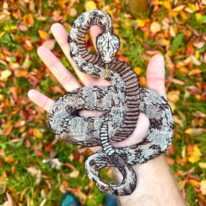 Peut inclure: Un patch brodé en forme de serpent, de couleur marron et blanc, avec un design détaillé. Le serpent est enroulé et a une apparence réaliste.