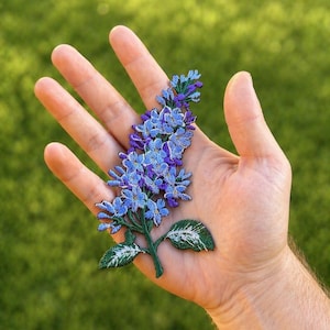 Könnte beinhalten: Gestickter Blumenaufnäher mit blauen und violetten Blüten, grünen Blättern und Stiel. Der Aufnäher ist ein detailliertes Design, das sich zum Aufnähen auf Kleidung oder Accessoires eignet. Der Aufnäher wird in einer Hand vor einem verschwommenen grünen Hintergrund gehalten.