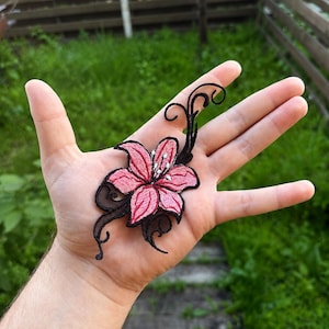 Puede incluir: Parche bordado con una flor de lirio rosa con detalles negros y remolinos. La flor tiene un diseño detallado con detalles blancos en el centro. El parche es probablemente para ropa o accesorios.