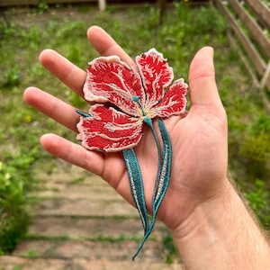 Op de afbeelding: Geborduurde rode bloemenpatch met gedetailleerde stiksels en blauwgroene stelen. De bloem heeft een delicate, getextureerde uitstraling, met een lichtroze omtrek op de bloemblaadjes. De patch is ontworpen om op stof te worden genaaid en voegt een decoratief bloemenelement toe.