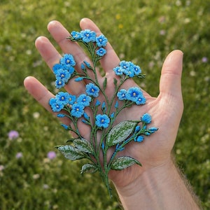 Könnte beinhalten: Eine detaillierte, gestickte Blumenapplikation mit leuchtend blauen Blüten, gelben Zentren und grünen Stielen und Blättern. Die Applikation wird in einer Hand gehalten, was die aufwendige Stickerei zeigt. Ideal zum Verzieren von Kleidung oder Bastelarbeiten.